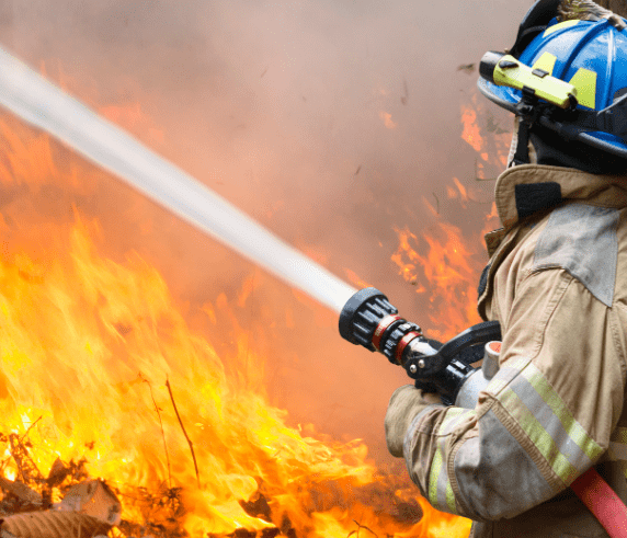 Fireman with a hose under his arm actively extinguishing a large wildfire in the background.