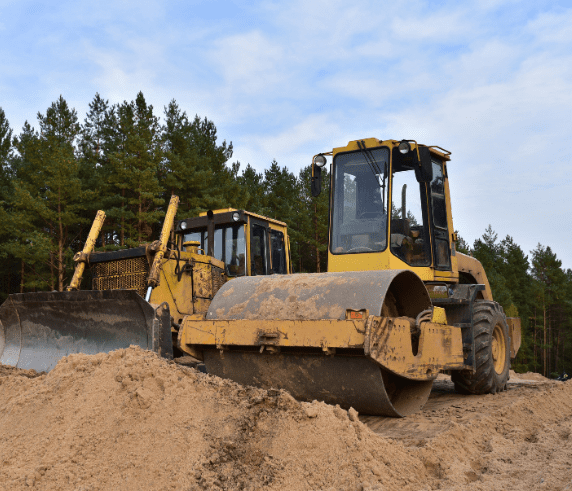 Student operating a roller during the Roller Ticket Perth / Roller Operator Ticket Perth course. Conduct Roller Operations. A yellow roller and a bulldozer on a sandy construction site with a forest in the background