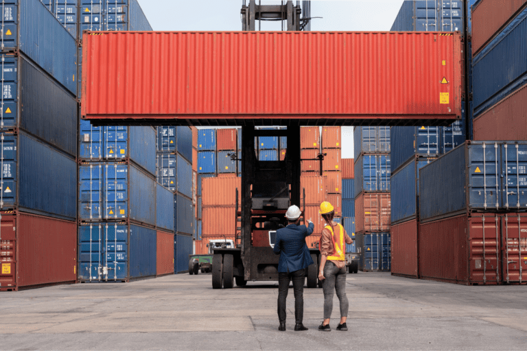 Reach stacker being operated at a docking yard moving a large red sea container being directed by two supervisors.