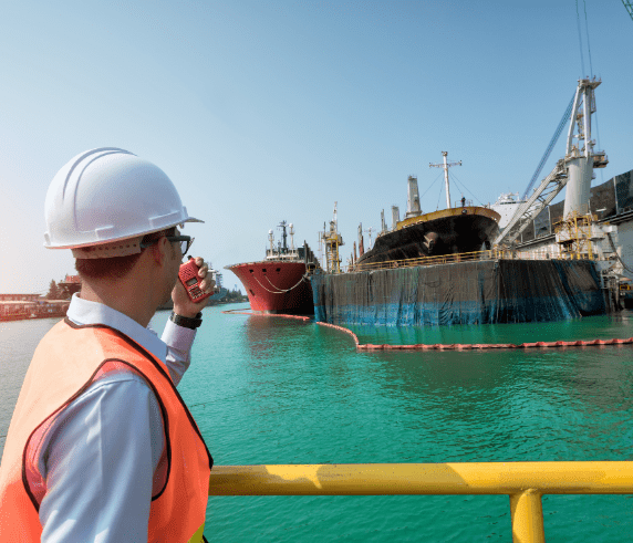 An individual in orange safety coveralls holding a clipboard and writing or reviewing notes in an industrial setting, possibly a factory or plant, with various pipes and valves in the background. The person’s face is obscured by a pixelated block for privacy, and there are bright yellow elements, likely safety barriers or parts of machinery, that stand out due to their color.