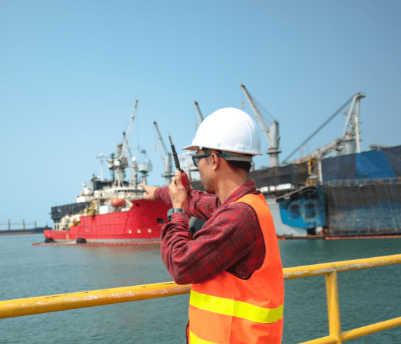 A person in safety gear standing on an offshore platform, looking towards a large red and black ship with cranes docked near the platform, under clear skies, indicating a maritime industrial setting or offshore construction and maintenance activities.