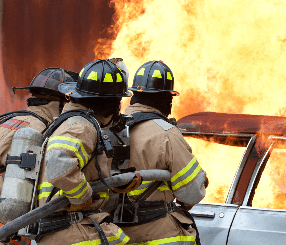Image depicts three firefighters in full gear, including helmets and oxygen tanks, standing near a vehicle engulfed in flames. The intense orange and yellow fire is blazing behind them, indicating an active and dangerous firefighting scene. The focus on the firefighters suggests their role in emergency response and the importance of fire safety measures.