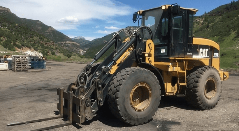 A Caterpillar integrated tool carrier parked on a gravel lot with mountainous scenery in the background. The machine features a large front-end loader with a fork attachment, robust tires, and a distinctive yellow and black color scheme