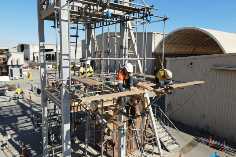 Scaffolding workers undergoing practical training at a Saferight facility. The workers are wearing safety harnesses and helmets while performing tasks on scaffolding at elevated heights. The background shows the training area with equipment and a warehouse in the distance