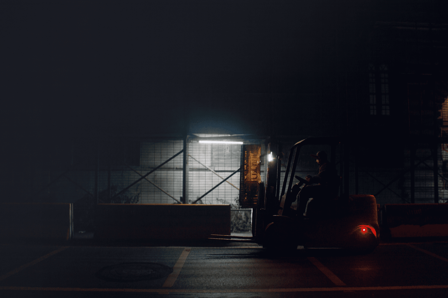 A forklift being operated at night under a dim streetlight, with its headlights and taillights illuminating the dark urban street and scaffolding in the background.